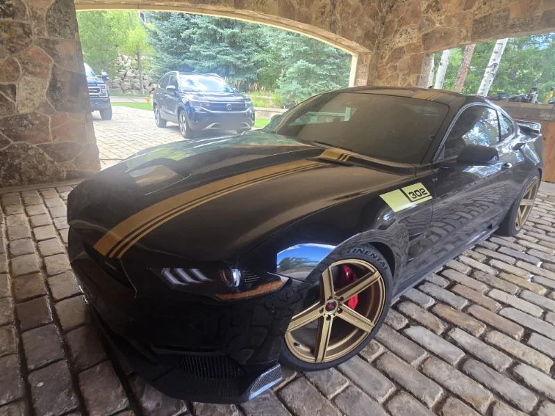 Black Ford Mustang with gold stripes and gold wheels under a stone carport